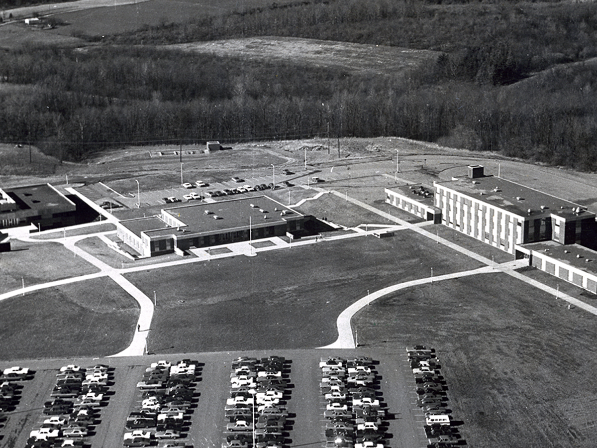 Black-and-white aerial photograph of Penn State Fayette, The Eberly Campus, showing several connected academic buildings, parking lots filled with cars, and open fields and woods in the background.