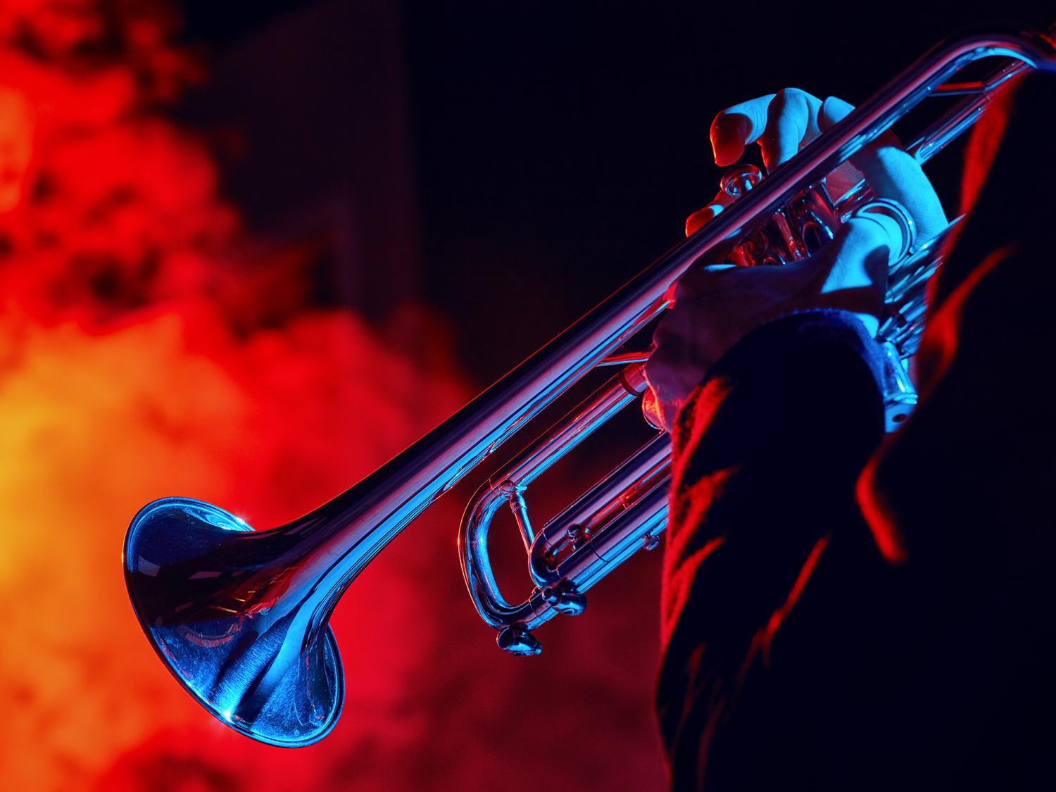 Close-up of a person playing a trumpet, illuminated by dramatic blue and red stage lighting with smoky background effects.