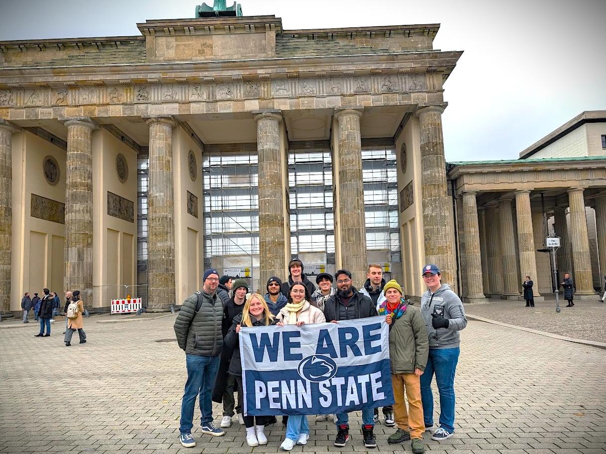 A group of students posing with a "We Are Penn State" flag in front of a building
