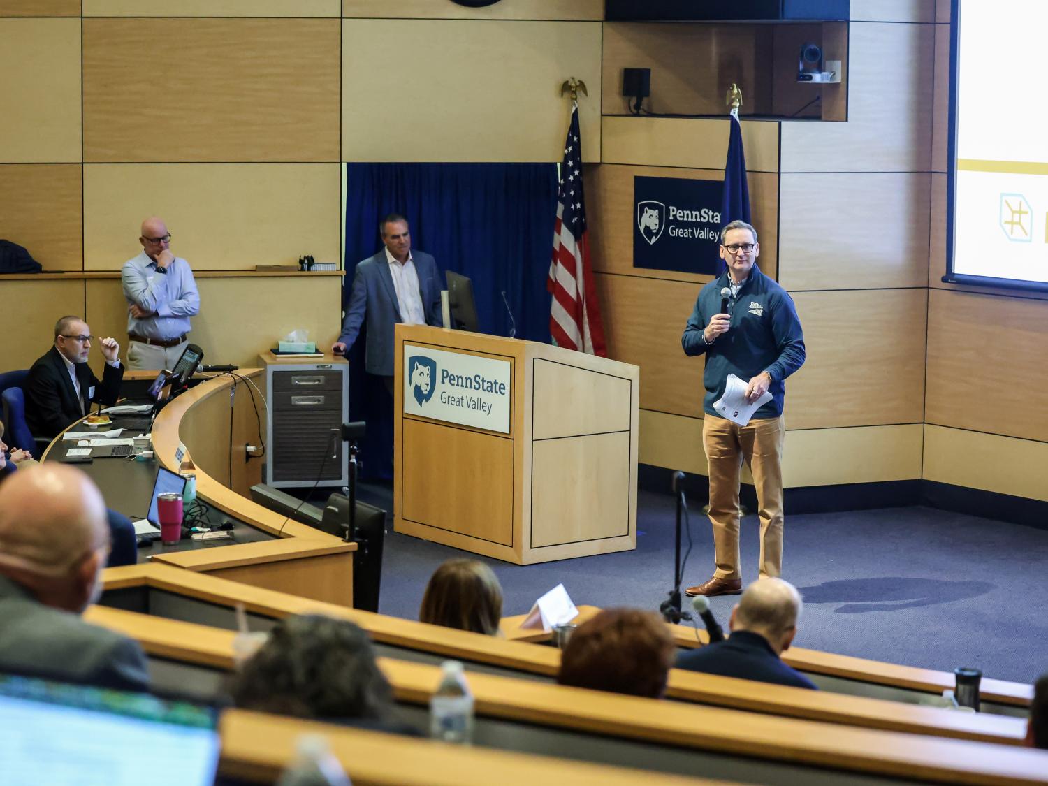 A man stands on a stage, speaking into a microphone, to a crowd of people seated around him.