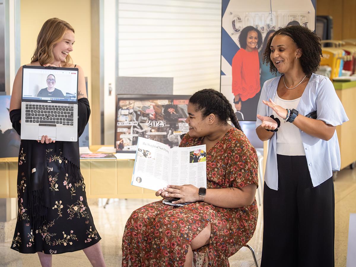 Three women childhood cancer survivors smile during an art exhibit at Penn State Health Golisano Children’s Hospital. The woman on the left is standing and holding a laptop that displays an image of a smiling woman. The woman in the middle is sitting and holding a magazine open, and the woman on the right is clapping. 