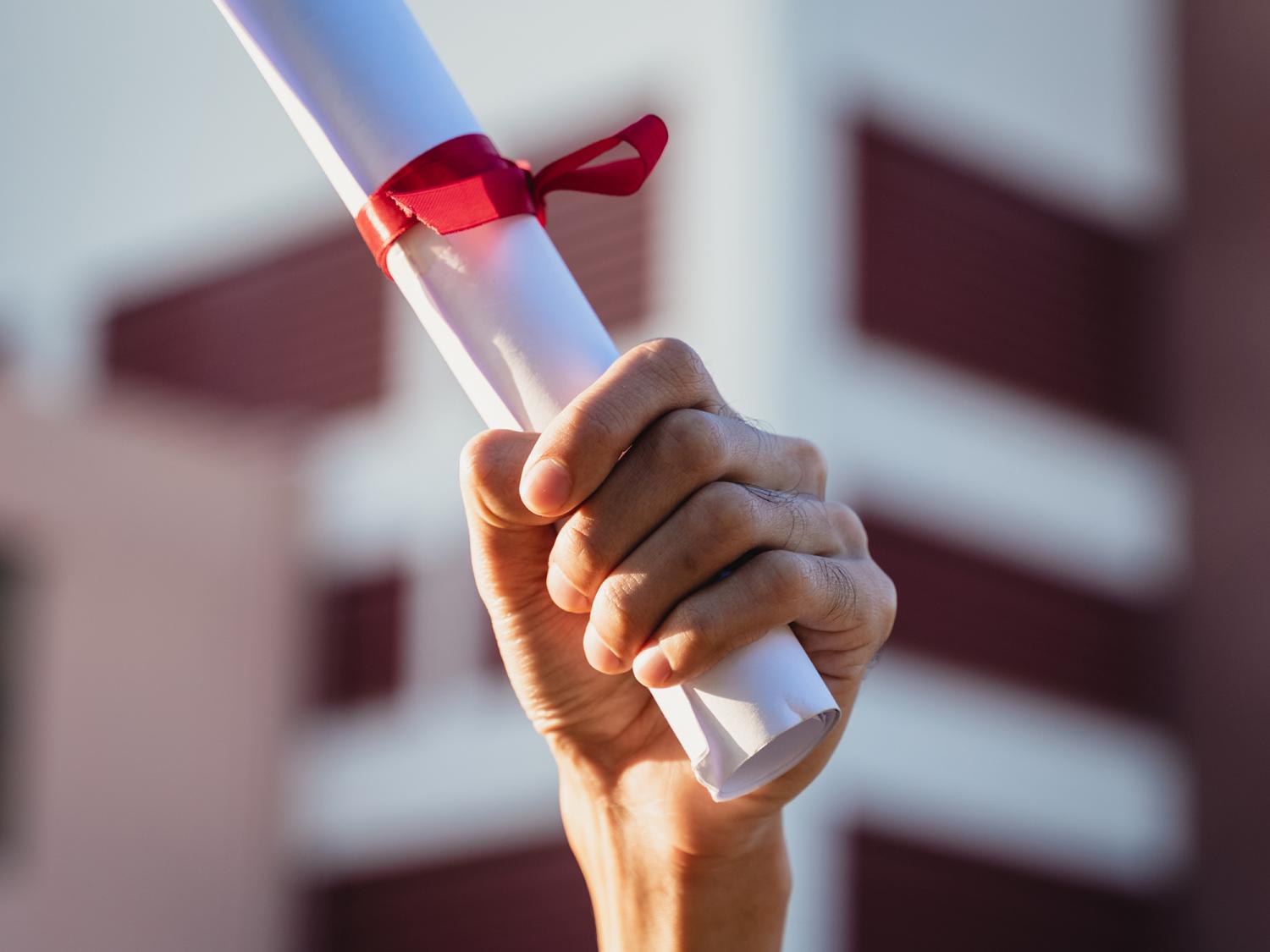 Hand holding up a rolled diploma tied with a red ribbon against the backdrop of a building in soft sunlight.