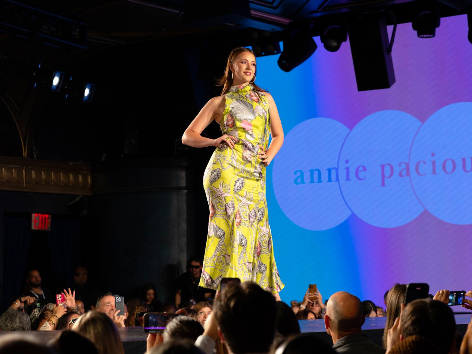Isabelle Bartruff walks confidently down the runway wearing a bright patterned dress during a fashion show before an audience.