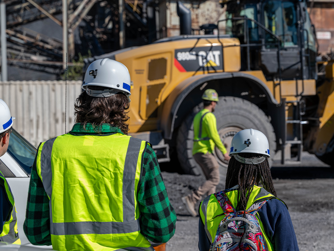 Students in safety gear at a construction site with a large yellow vehicle in the background.