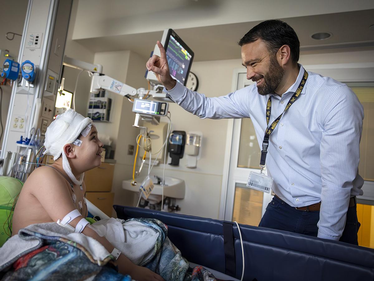 Brent O'Neill, right, wearing a shirt and tie, raises a finger and smiles at a boy in a hospital bed. The boy is looking up at him. The boy’s head is covered in bandages, he has an IV in his arm and has a blanket on him. Behind them are a monitor and medical equipment.