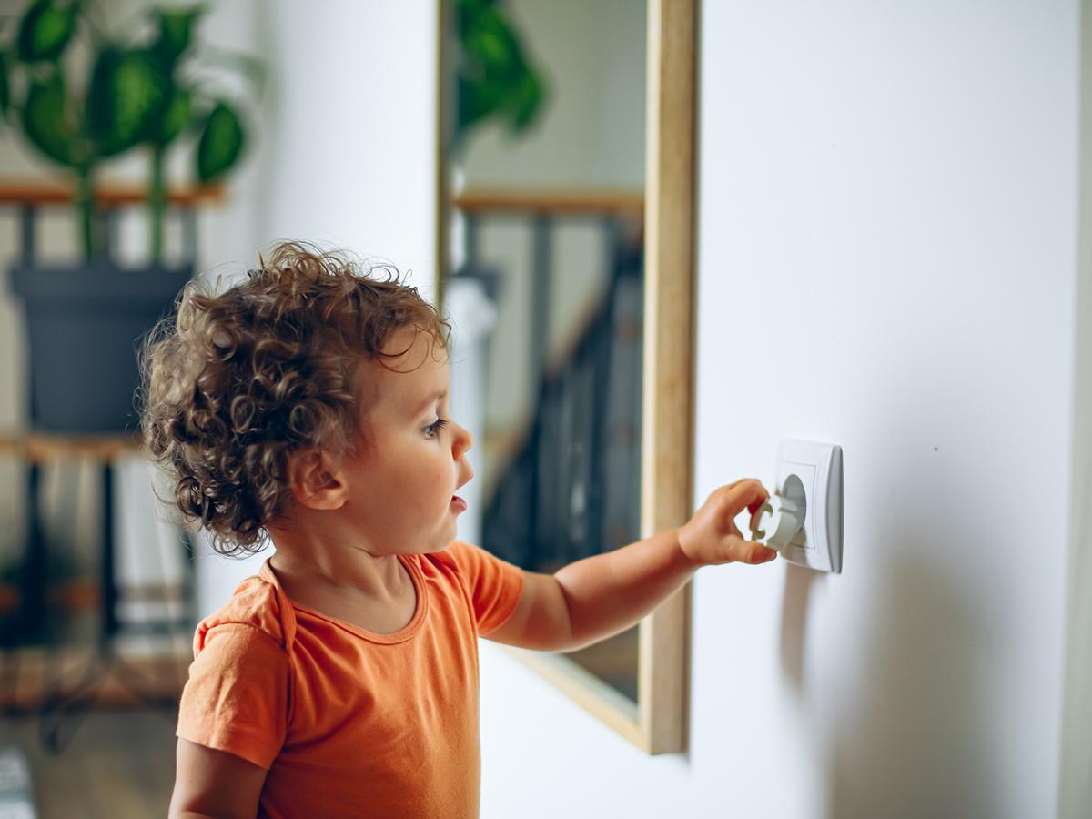 Toddler reaches into electrical outlet on wall with her hand.