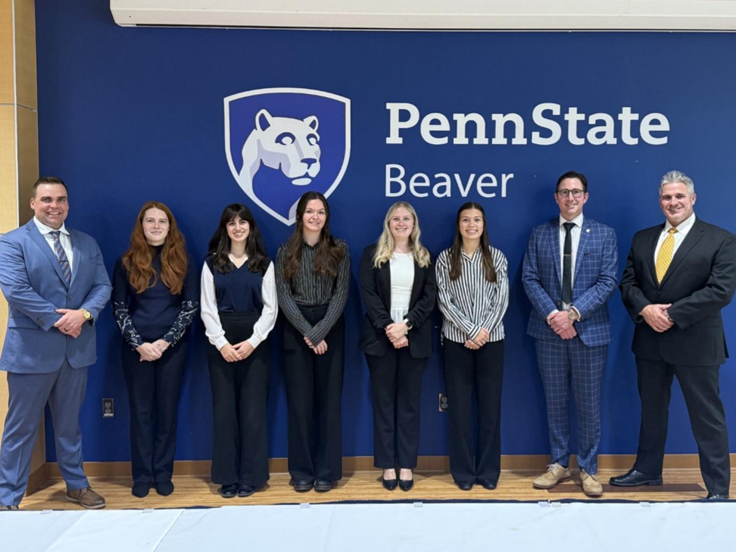 A group of eight people in business attire pose in front of a blue wall with the Penn State Beaver logo and lettering.