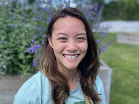 A headshot of a woman in a t-shirt outside with long brown hair and smiling