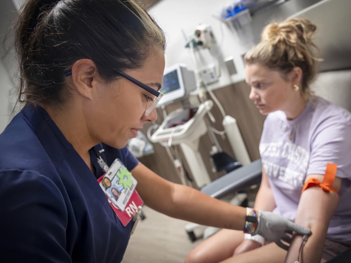 A nurse wearing navy scrubs and glasses draws blood from a patient seated in a medical chair.