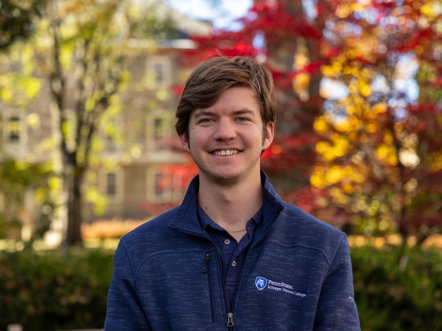 Morgan Overman stands in front of green, red, and yellow trees while wearing a navy Schreyer Honors College quarter-zip at Penn State University Park.