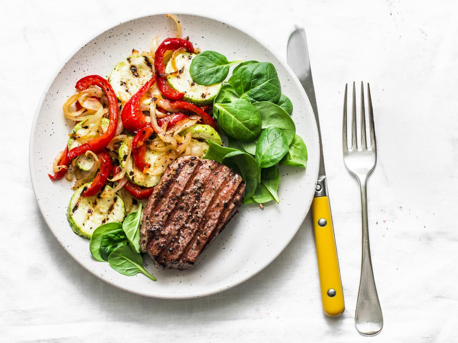 small piece of steak on a plate with raw spinach and roasted vegetables