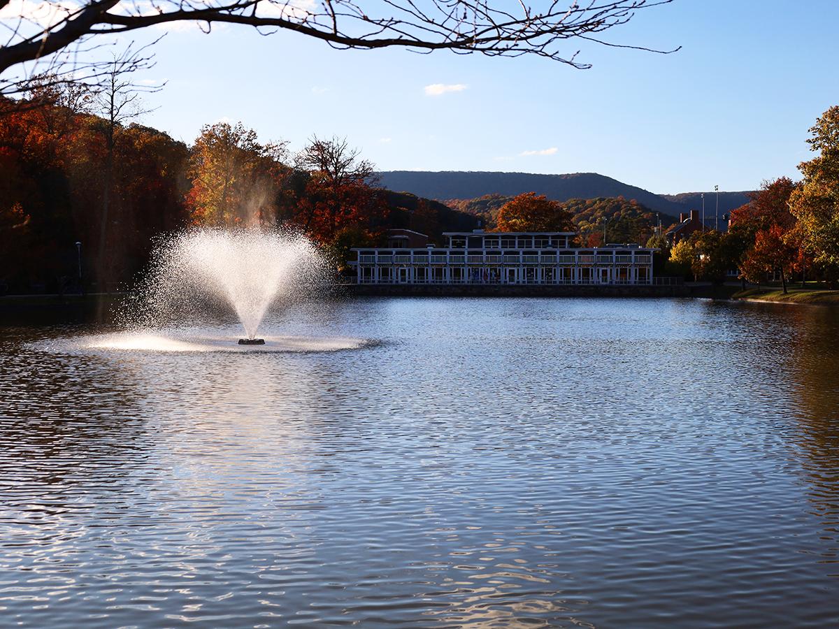 Fountain sprays water in a calm campus pond surrounded by colorful autumn trees with a modern building and hills in the background.