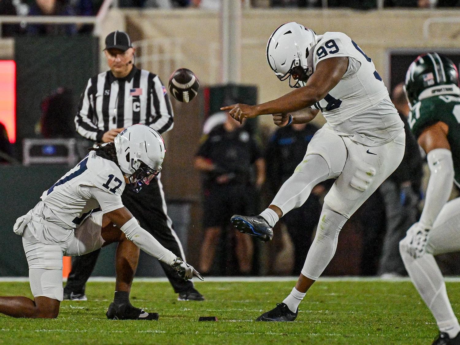 A photo of No. 99 Gabe Nwosu, in Penn State's away white uniforms, kicks off as No. 17 holds the ball and a referee looks on in the background.