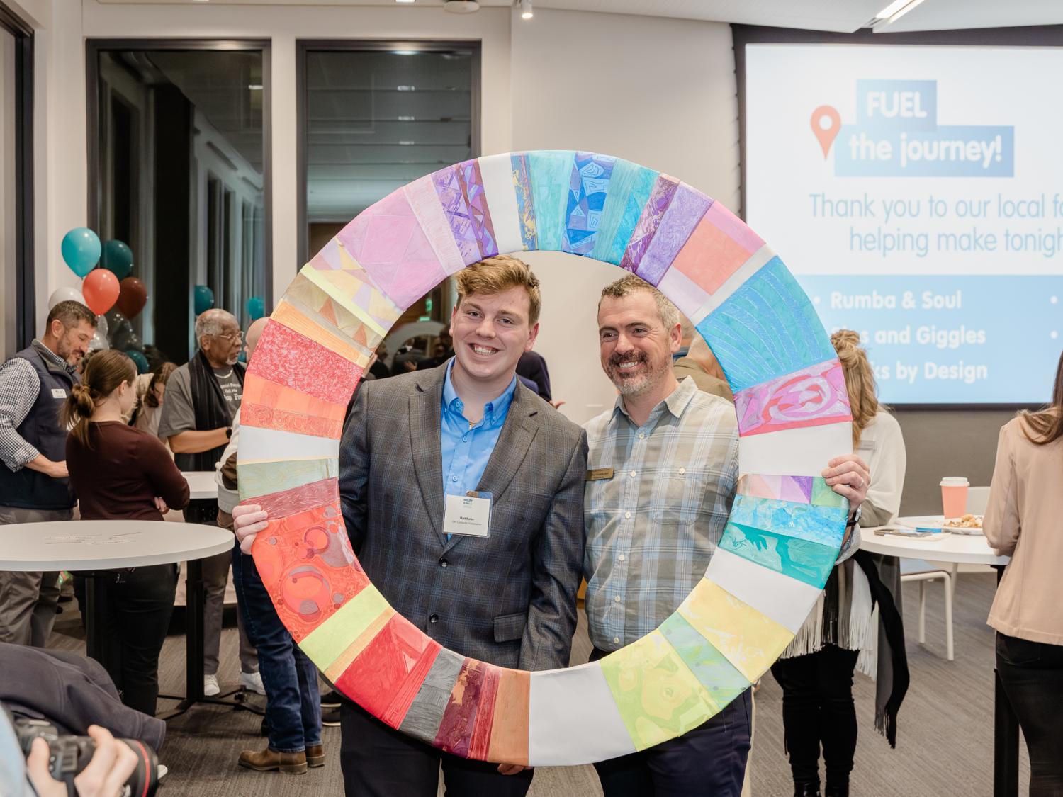 Two individuals pose holding up a large, GEW-themed colorful circle to frame themselves