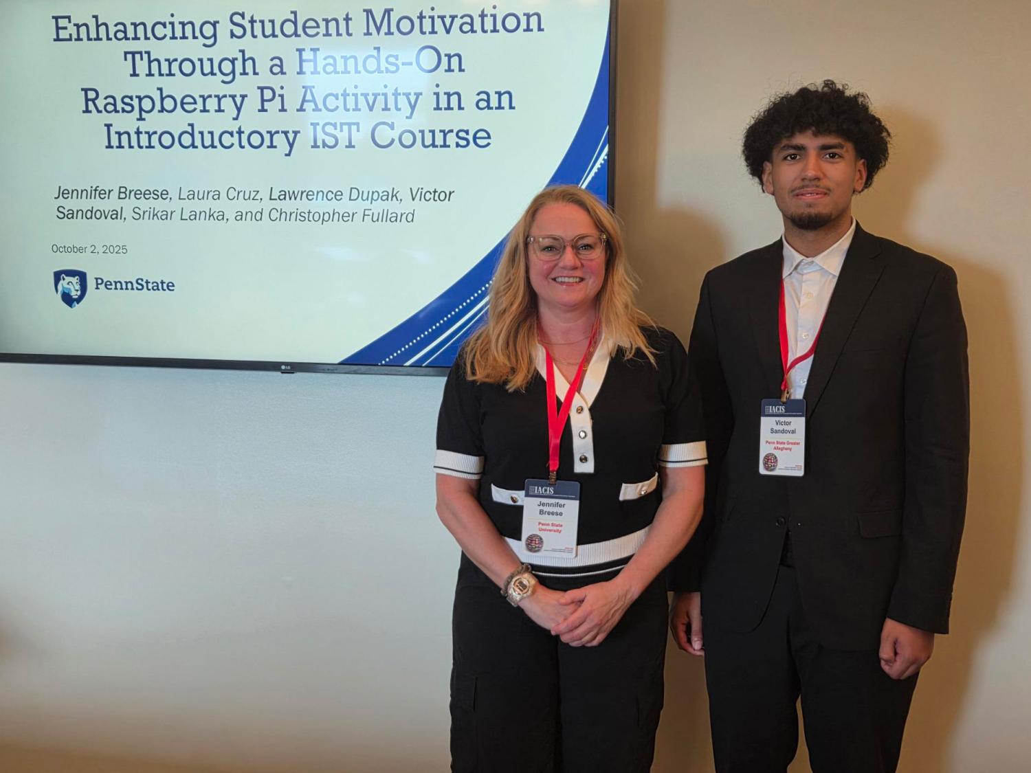 Two people stand side by side in front of a presentation screen displaying the title of a study on enhancing student motivation through a hands-on Raspberry Pi activity. Jennifer Breese is on the left, wearing glasses and a black dress with white trim. Victor Sandoval is on the right, wearing a black suit and white shirt. Both wear conference badges and face the camera, smiling.