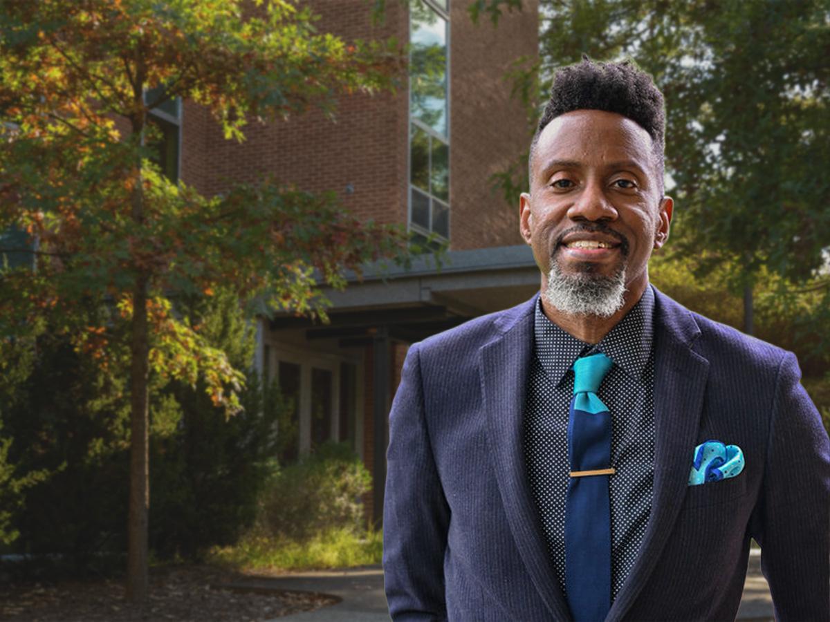 A man wearing a navy suit jacket, patterned shirt and teal tie stands outdoors in front of a Penn State Greater Allegheny building, with trees and fall foliage behind him.