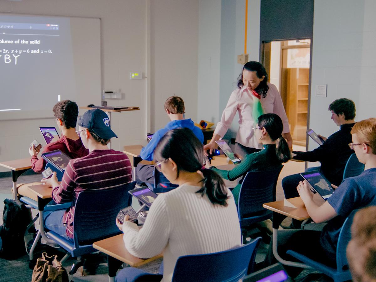Kuei-Nuan Lin stands beside seated students in a classroom at Penn State Greater Allegheny, assisting them as they work on tablets during a mathematics lesson.