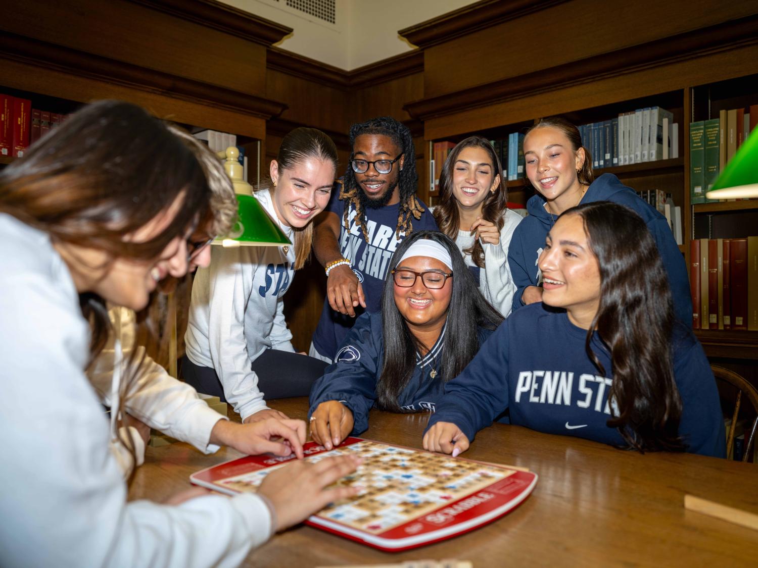 A group of students sits and stands around a wooden table in a library, leaning in toward a board game. They are smiling and interacting with the game pieces. Bookshelves line the background, and warm lighting from green-shaded lamps illuminates the scene.