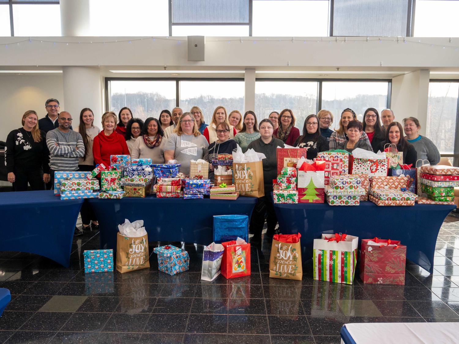 A group of more than 20 people stand behind tables laden with colorful, beautifully wrapped holiday gifts.