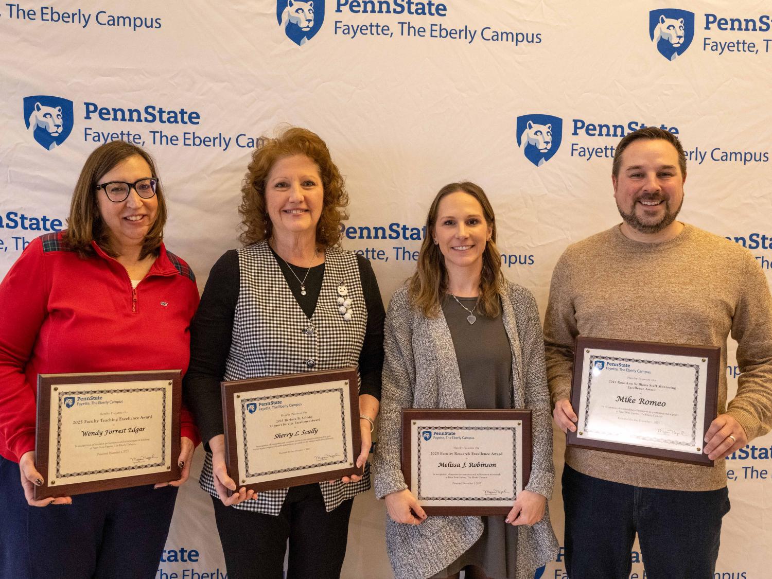 From left, Wendy Edgar, Sherry Scully, Melissa Robinson, and Mike Romeo stand in front of a Penn State Fayette, The Eberly Campus banner holding framed awards and smiling.