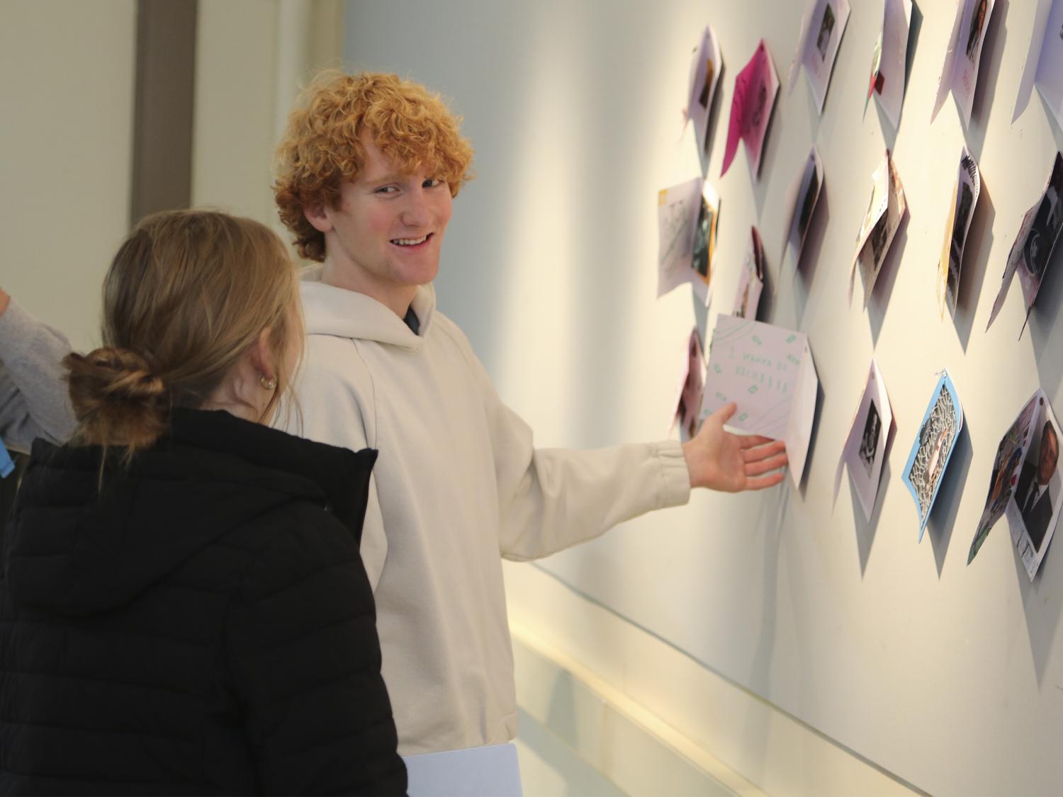 Two students look at each other and smile as one gestures towards art displays hanging on a wall.
