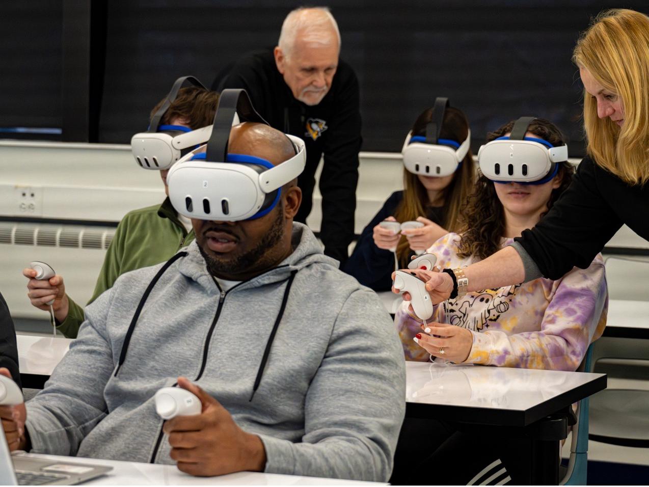 Students wearing VR headsets sit at desks in a classroom, holding controllers as an instructor assists one of them. Another instructor observes from behind.