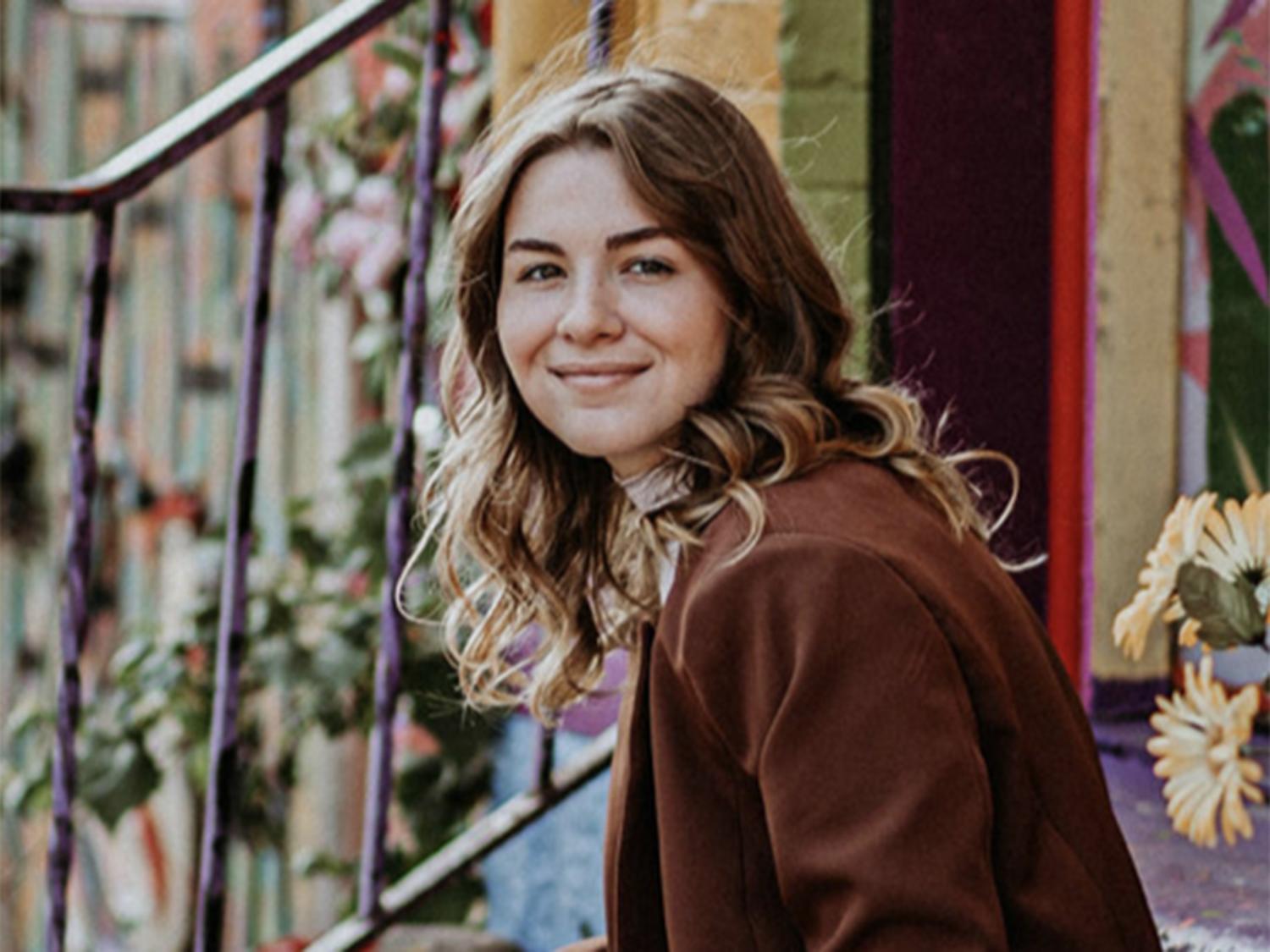 Girl with dark shoulder-length hair wearing a coat while sitting on a stoop (a small set of stairs).
