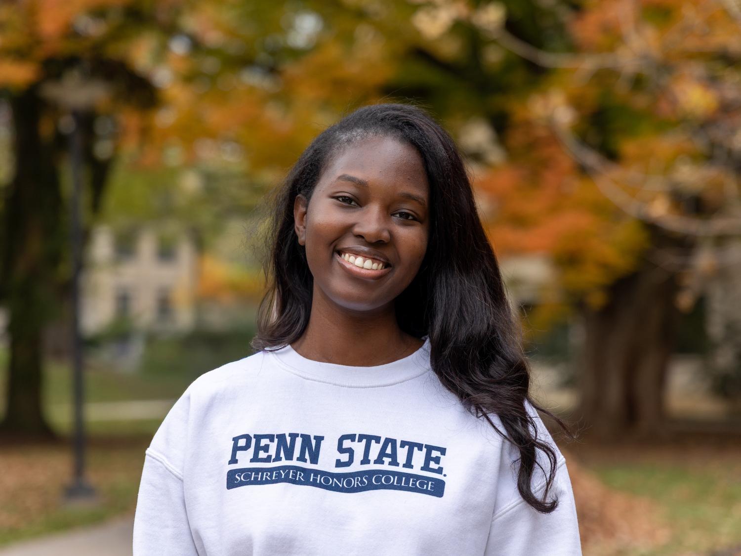 Penn State student Fatoumata Doumbia stands in a white Schreyer Honors College sweatshirt in front of trees at Penn State University Park in autumn.