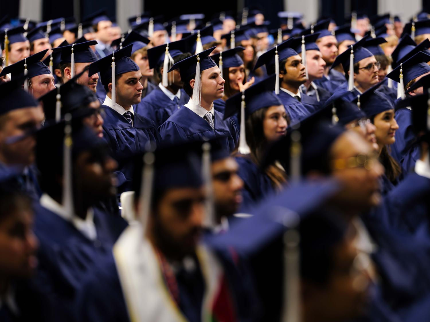 Rows of graduates seated indoors at a commencement ceremony, wearing navy blue caps and gowns with white tassels, facing forward and listening attentively during the program.