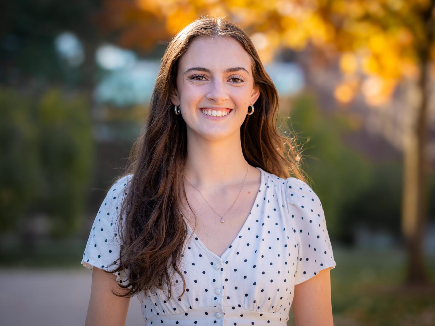 smiling person with long dark hair against backdrop of sunlit trees