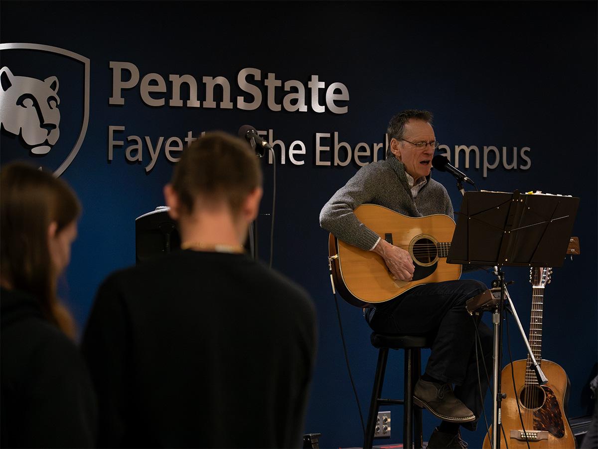 Chuck Cantalamessa plays guitar and sings on stage in front of a Penn State Fayette, The Eberly Campus backdrop.