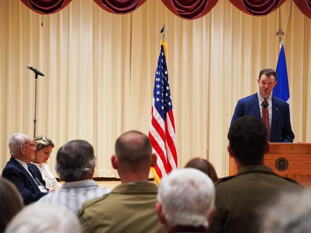 Jared Frederick at a podium addresses an audience in an auditorium, with U.S. flag and John Homan looking on.