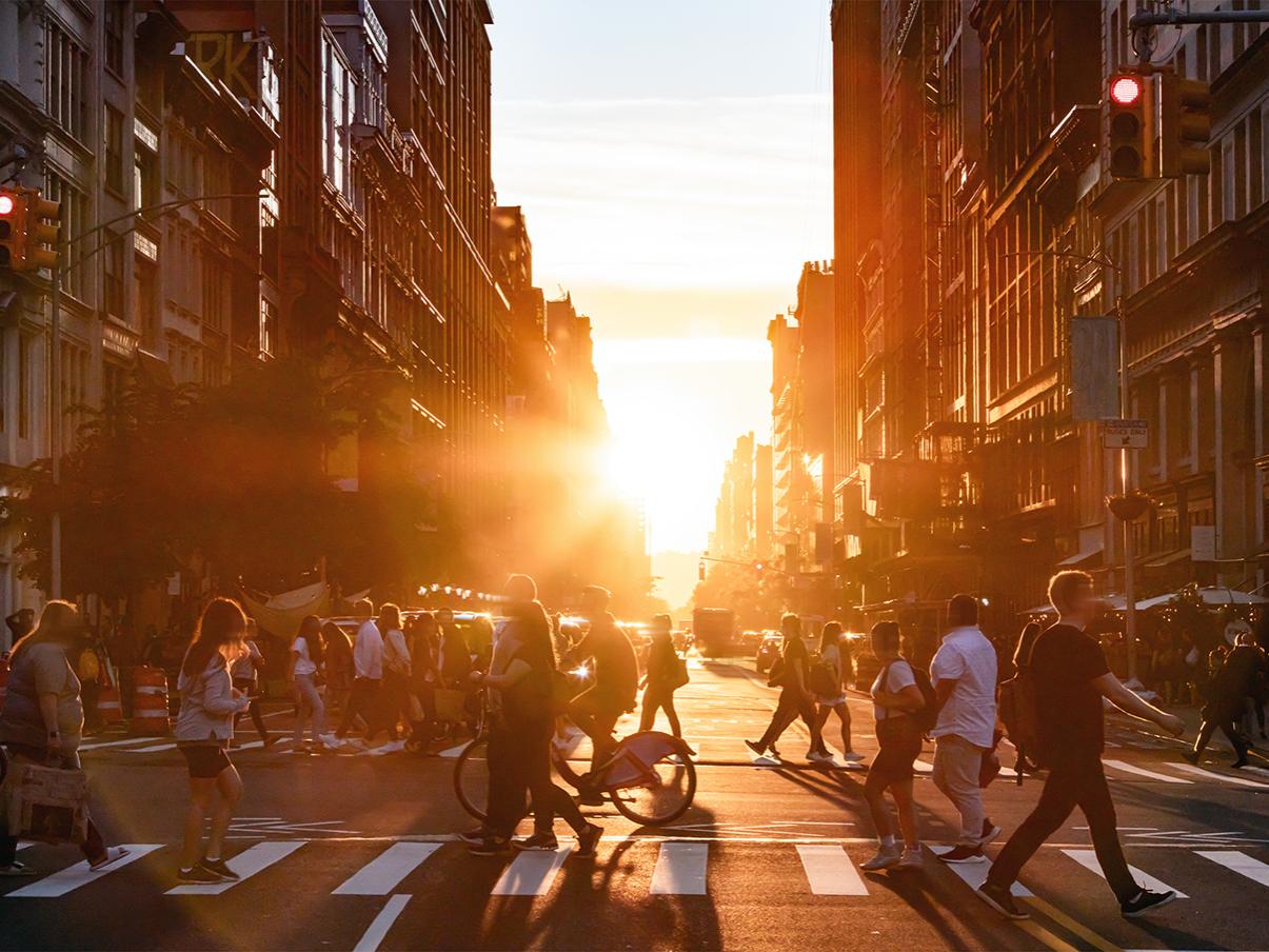 People walk through a busy intersection in New York City on a summer day with sunset flare between the background buildings