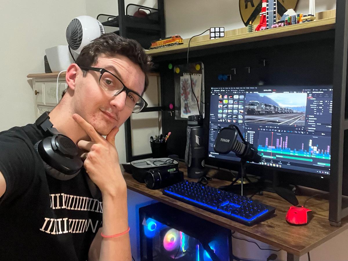Kian Woods posing at a desk with a computer editing video footage, surrounded by tech gear, lights, and model trains on shelves.