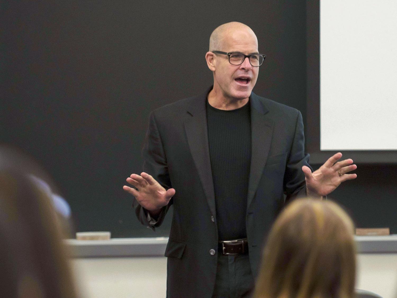 Bald man with glasses in a brown suit coat and black shirt gestures with his hands in front of a class