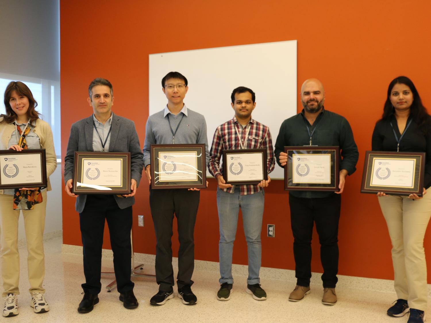 Six people posing in front of an orange wall with their award plaques.
