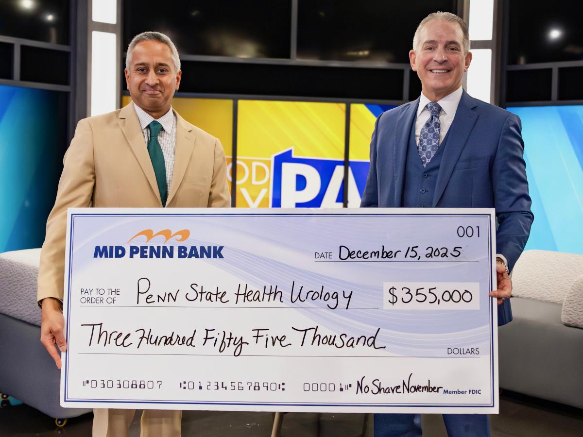 Two men in professional business attire smile as they pose for a photo with an oversized check in the amount of $355,000 made out from Mid Penn Bank to Penn State Health Urology. A TV station set is in the background.