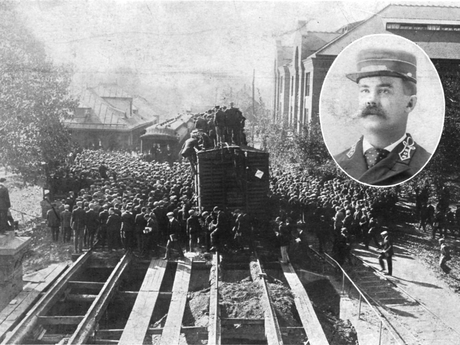 An historical image of train cars at a station with at least a hundred people around and on top of it