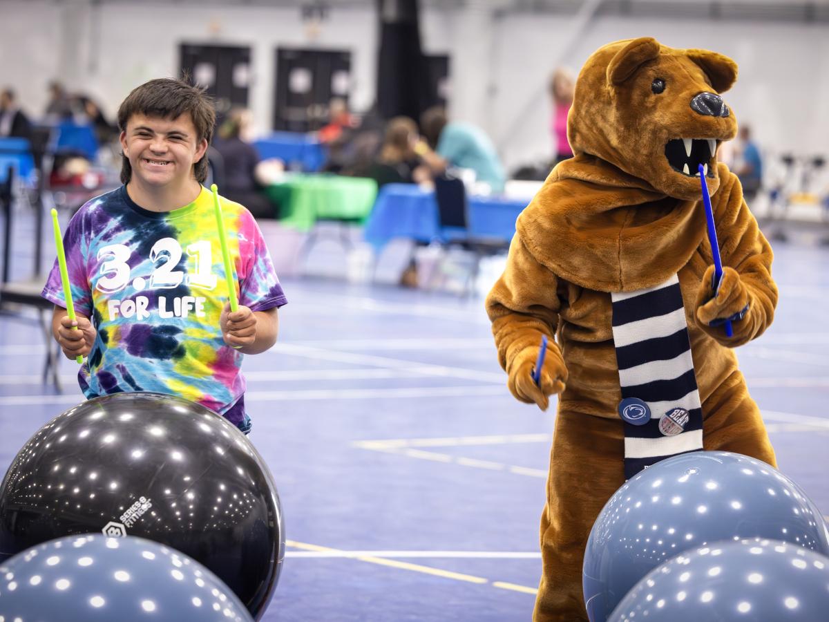 A teen boy and the Nittany Lion mascot use drumsticks to drum on exercise balls in a large gym at Penn State Health RecFest 2025. The boy is wearing a tie-dye T-shirt with the words “3.21 for Life” on it. The Nittany Lion has a scarf.