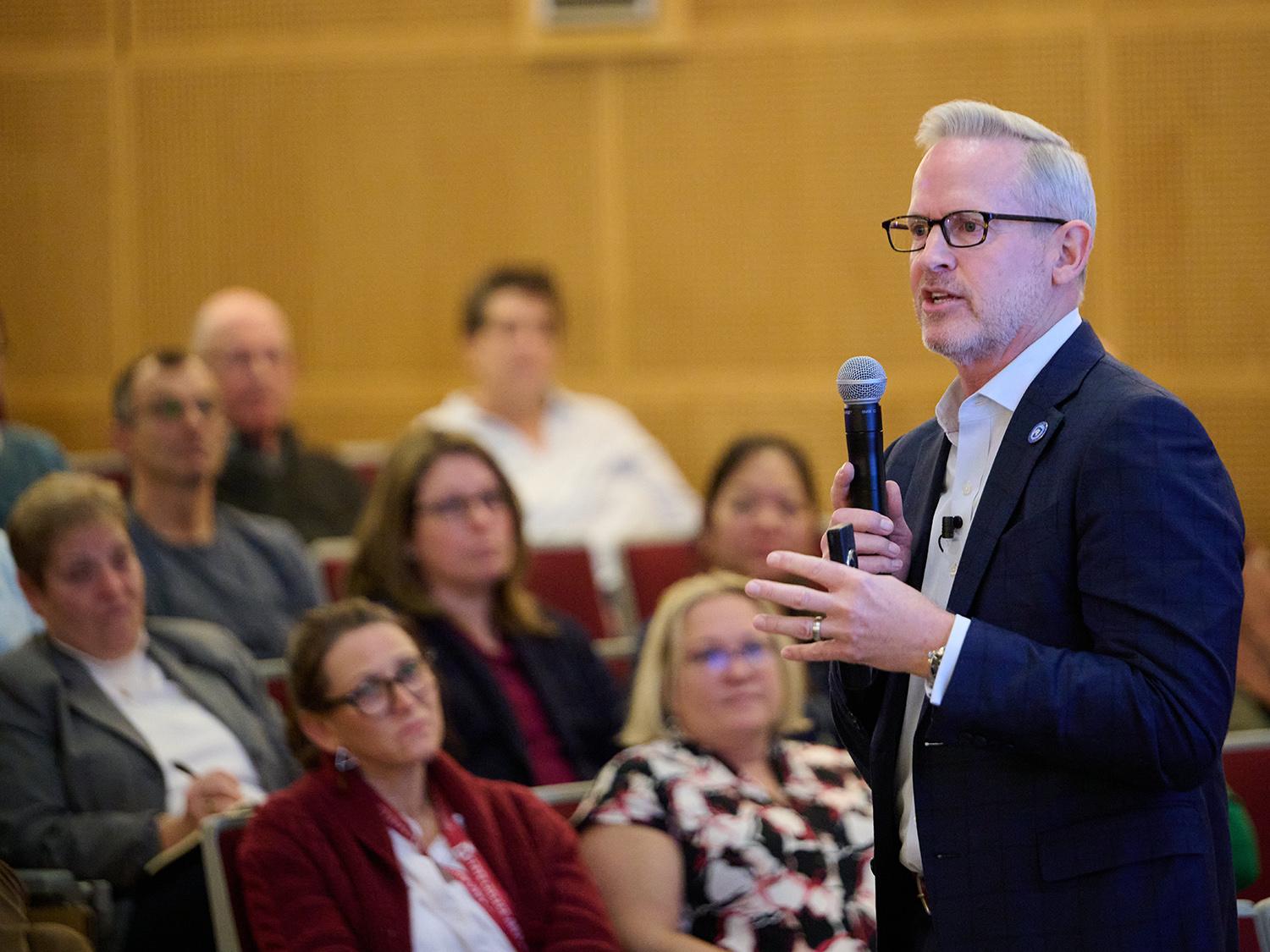 Smeal College of Business Dean Corey Phelps, dressed in a blue blazer and white shirt and wearing glasses, stands in an auditorium with a microphone in his hand.