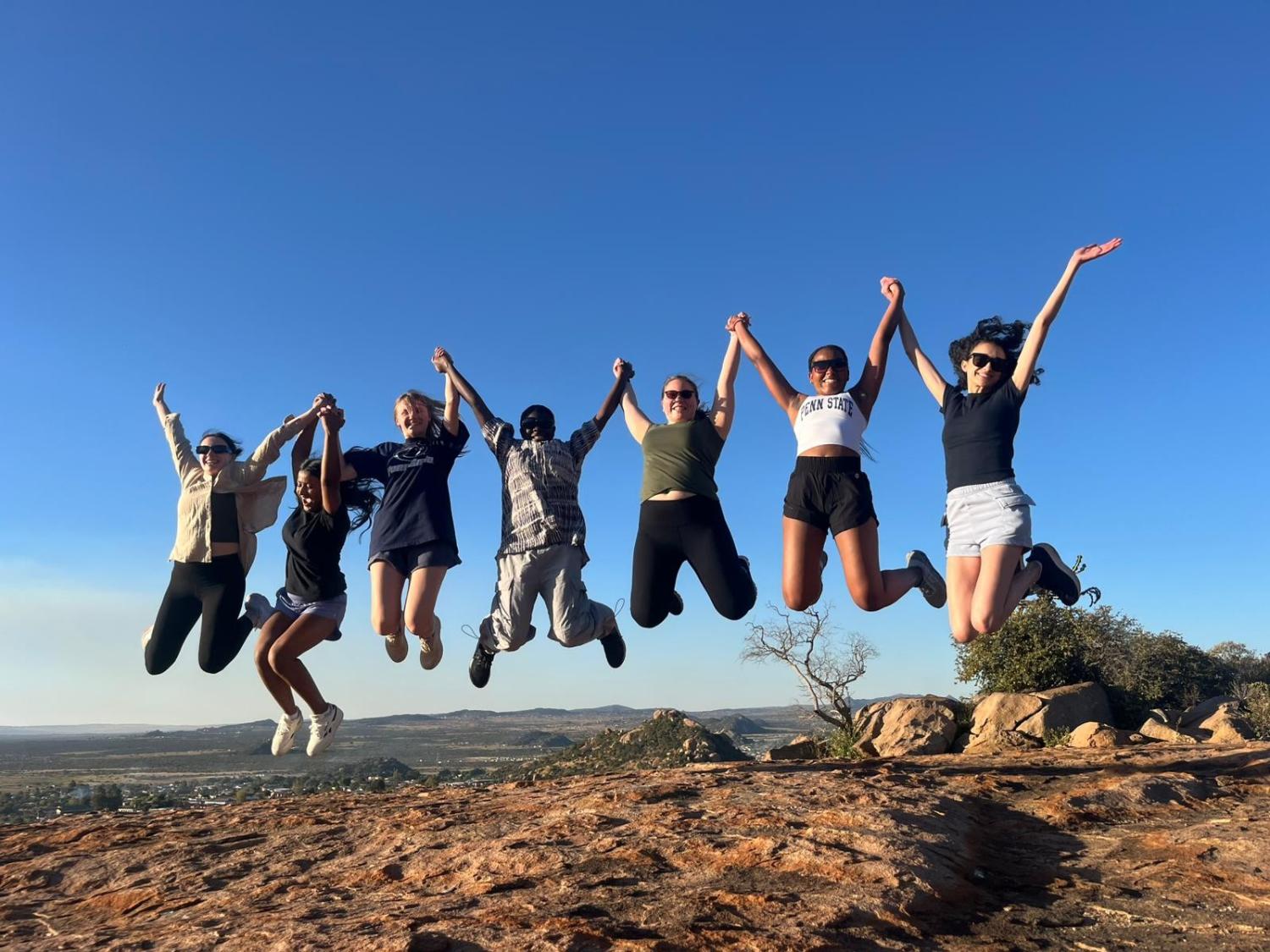 Seven Penn State students in front of South African landscape jump in the air while holding hands