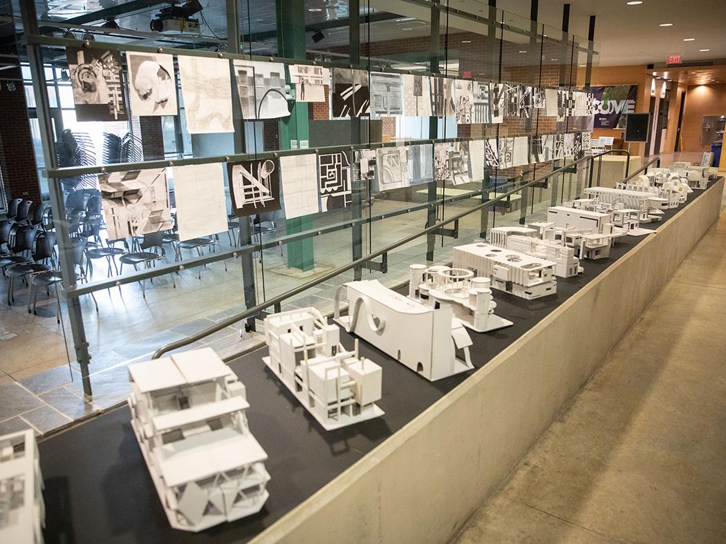Architectural models lined up on a black surface with plans hung over head.