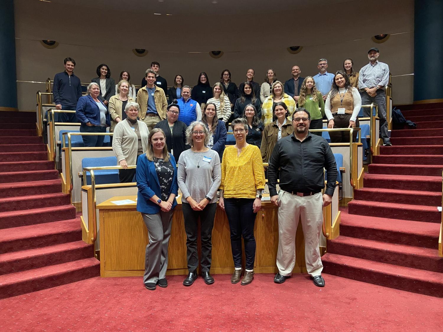 A group of 31 people stand in front of four rows of raised seating during the LCAP Town Hall Conference of Parties in Harrisburg
