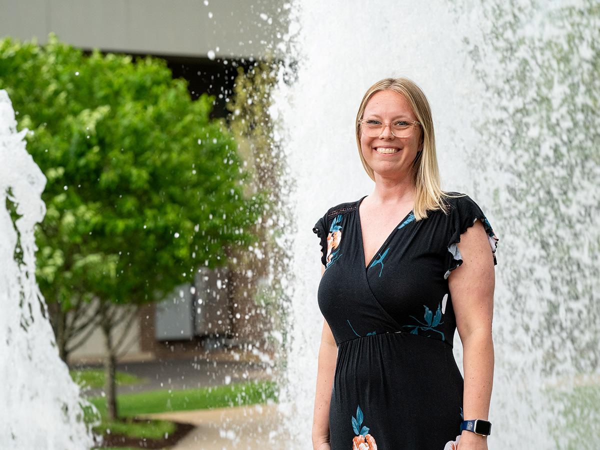 Amanda Collins stands smiling beside a fountain at Penn State Fayette, The Eberly Campus, with campus buildings and greenery in the background.