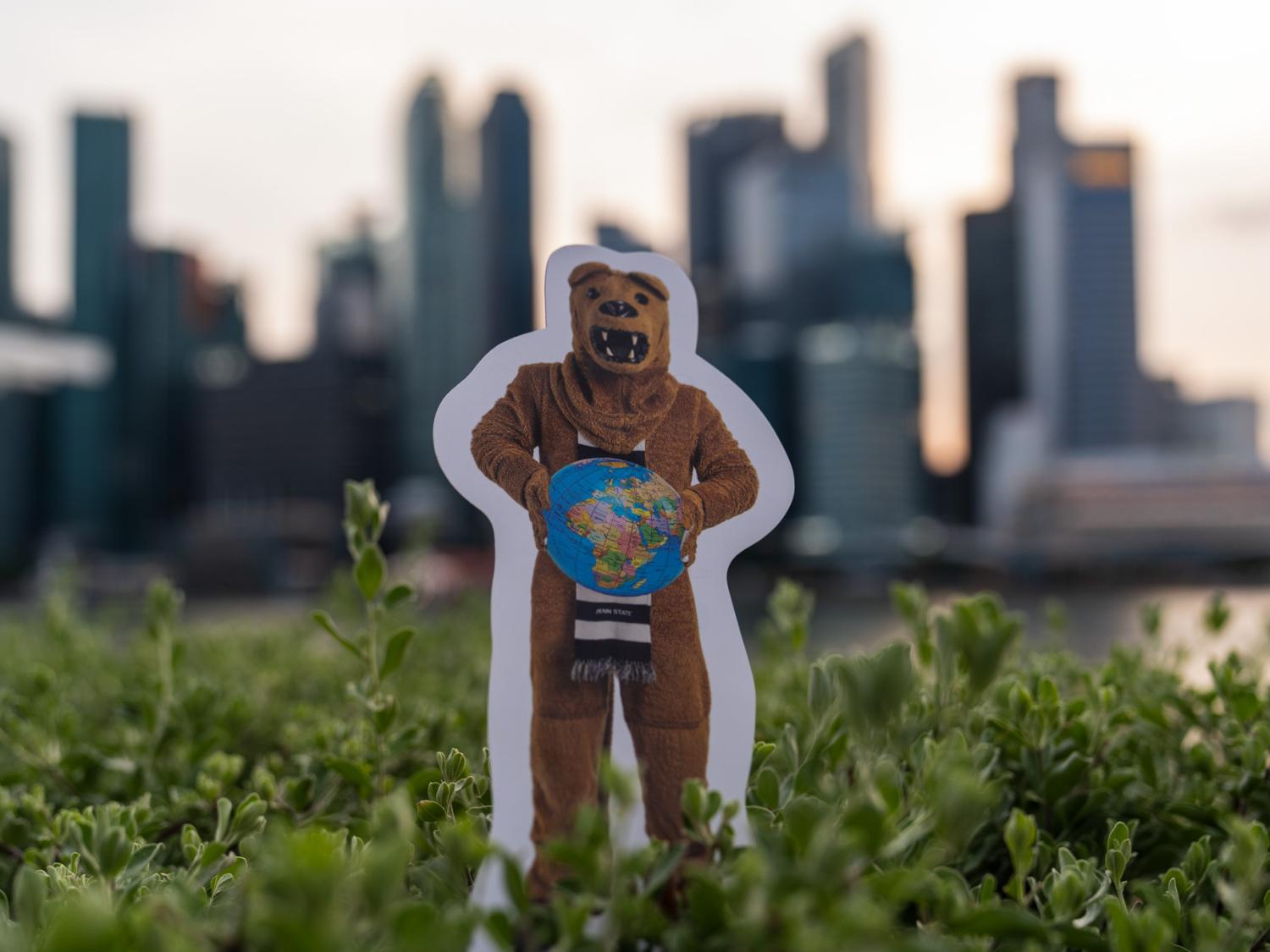 Picture of small Nittany Lion cutout holding a globe with a city skyline in the background.