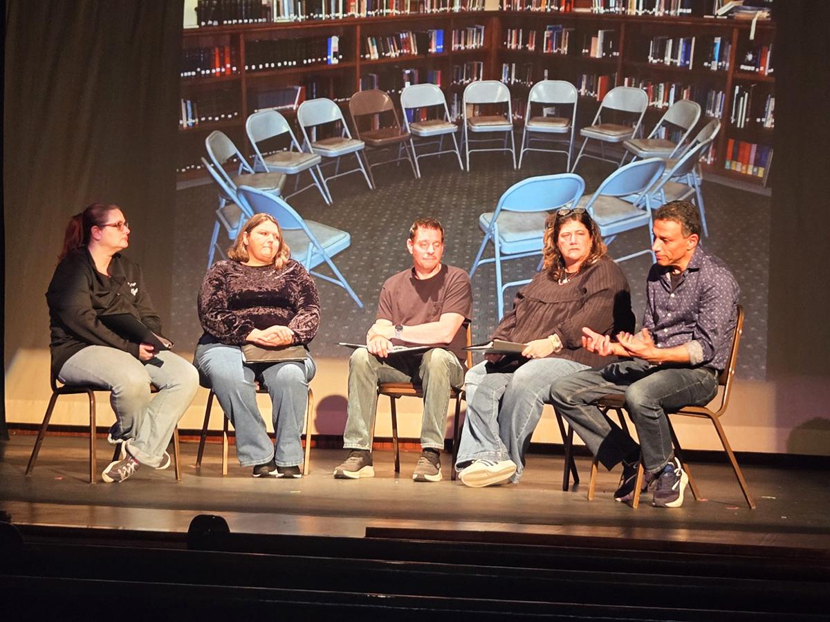 Five people seated on stage in a panel discussion, with a projected image of a circle of chairs in a library behind them.