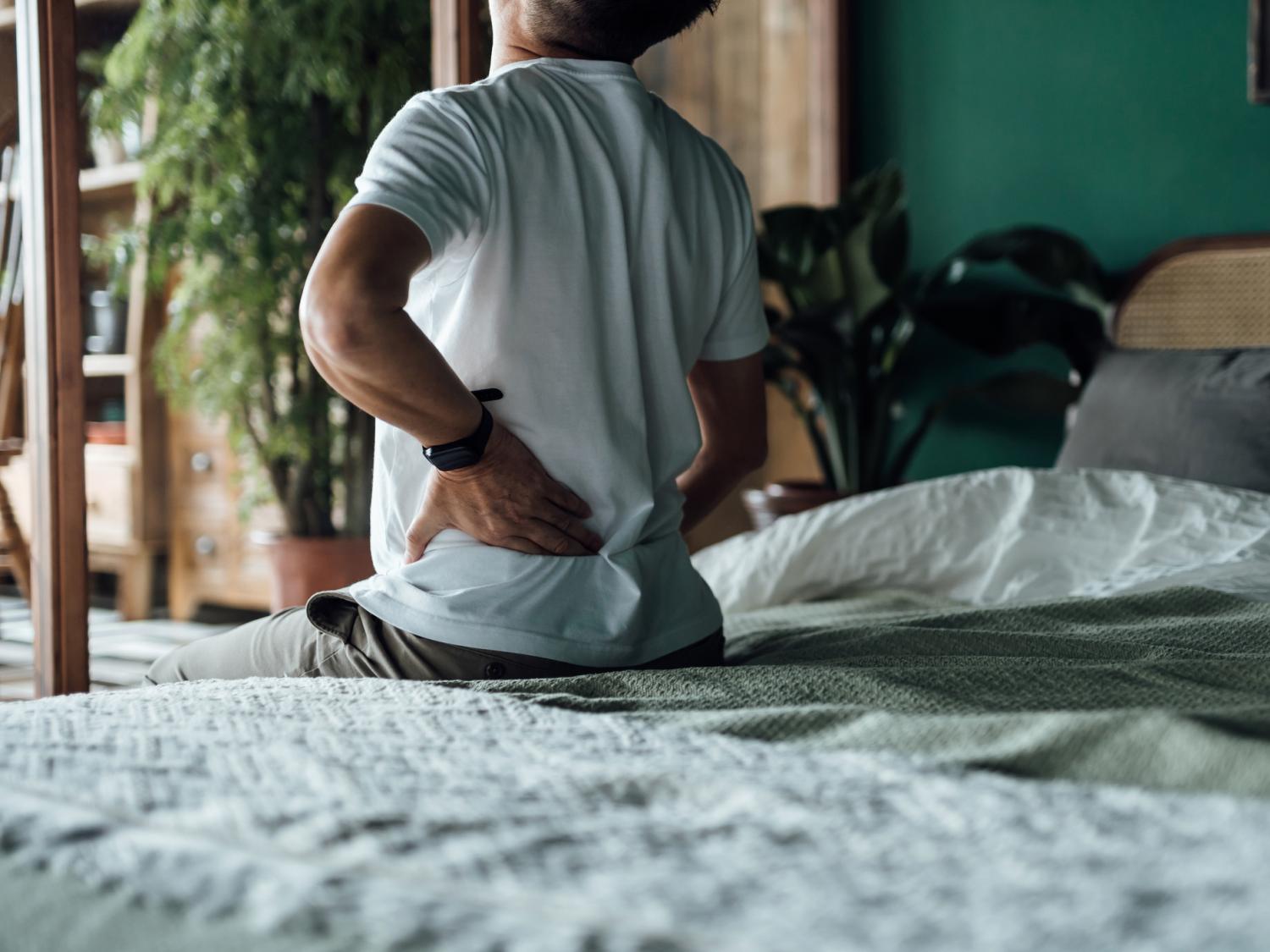 Older person sits on the edge of a bed with a hand on their back, indicating discomfort