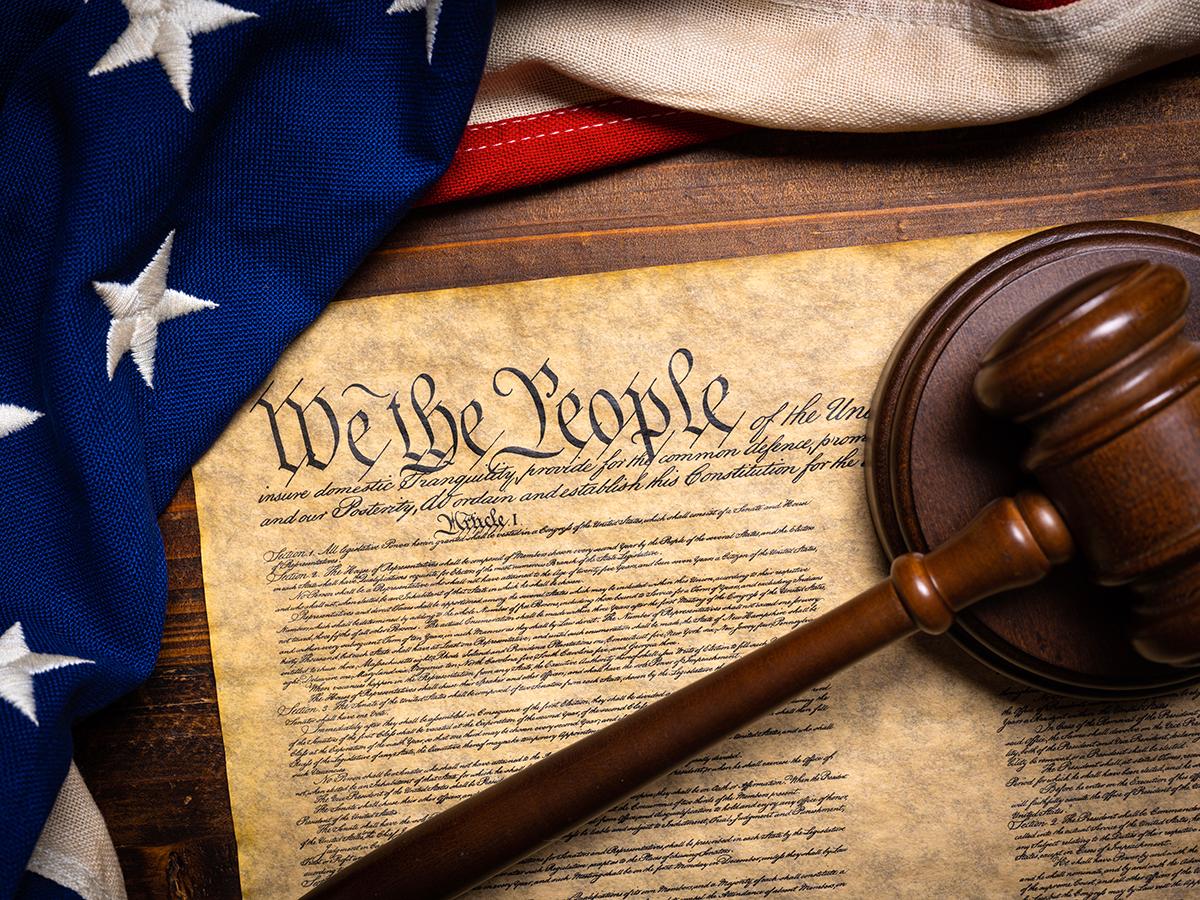 American flag draped beside a wooden gavel resting on the U.S. Constitution opened to the words WE THE PEOPLE.