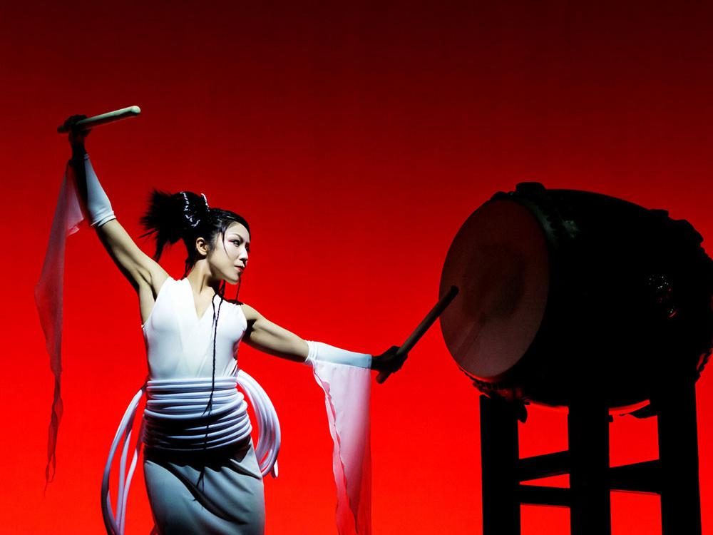 A Japanese woman holding large sticks and wearing a robe stands at a large drum.
