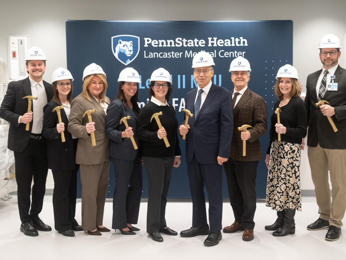 A group of hospital leaders and staff wearing white Penn State Health hard hats stand in a bright clinical room, each holding a gold ceremonial hammer. They pose smiling in front of a Penn State Health Lancaster Medical Center backdrop during a NICU groundbreaking celebration, with medical equipment visible on both sides of the room.